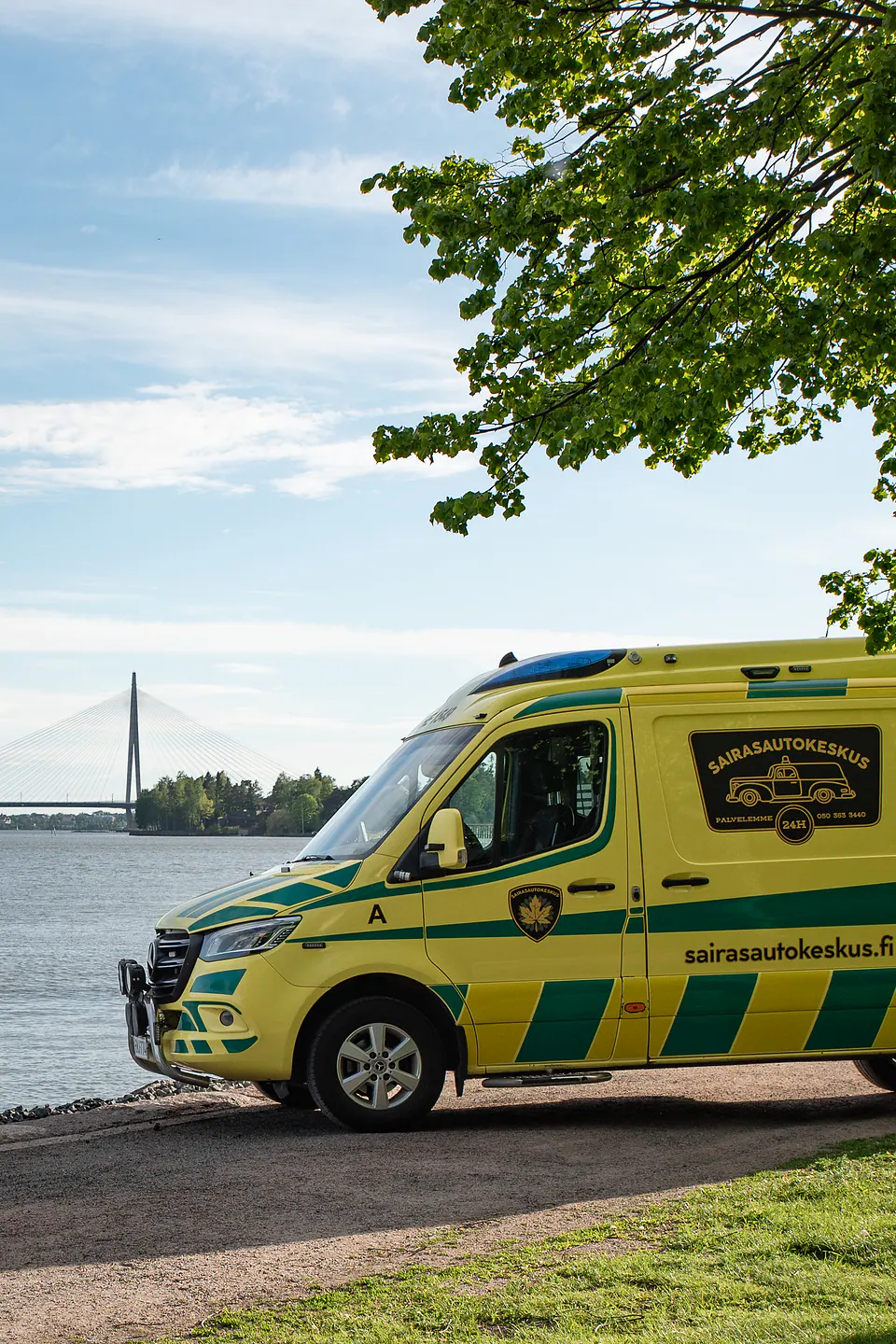 Ambulanssi rannassa Keltaisen ambulanssin etu- ja kylkiosa näkyy rantamaisemassa sillan ja lehvästön kehystämänä.