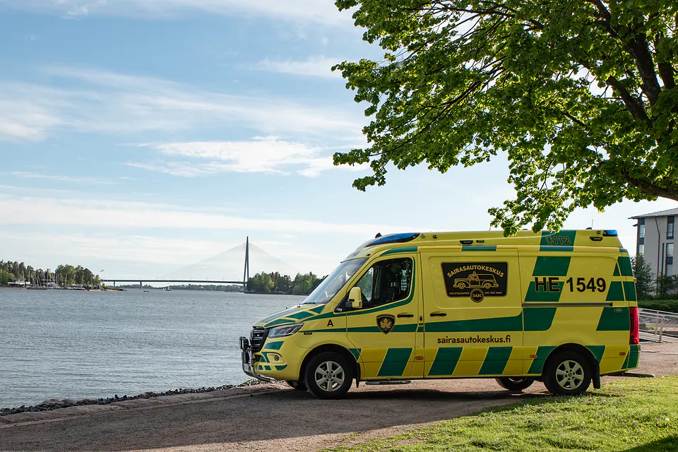 Kalusto rannassa Kokonainen keltainen ambulanssi on pysäköity rantareitille vesistön ja vinoköysisillan eteen.