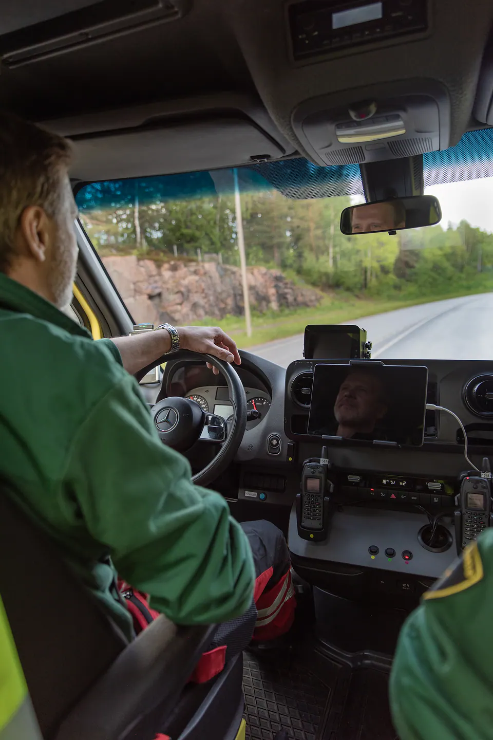 Tien päällä Työntekijä ajaa ambulanssia metsäisen tien varrella toisen työntekijän istuessa vieressä.