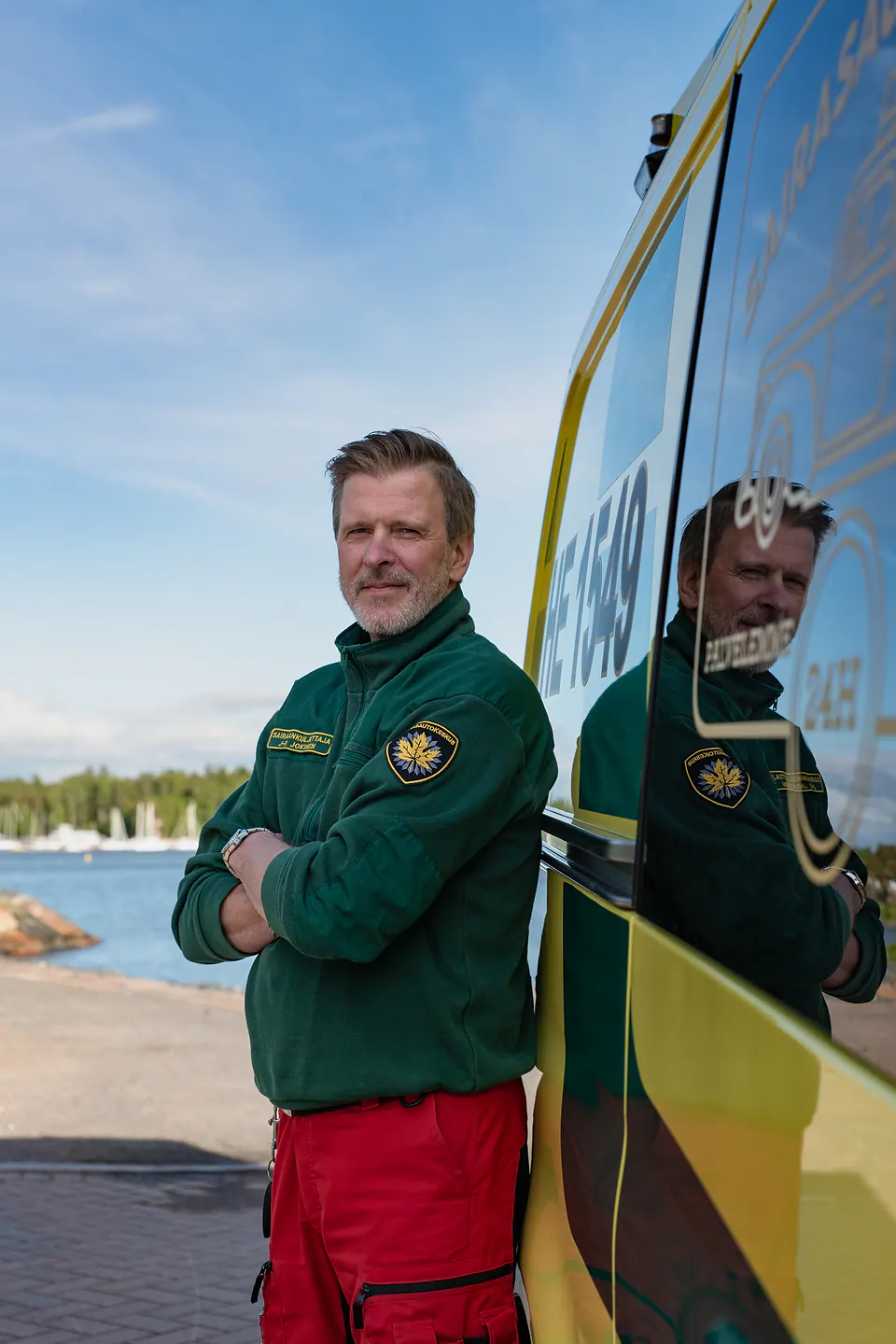 Ammattilainen työympäristössä Työntekijä seisoo kädet ristissä ambulanssin vierellä vihreässä työpaidassa, vesistö taustallaan.