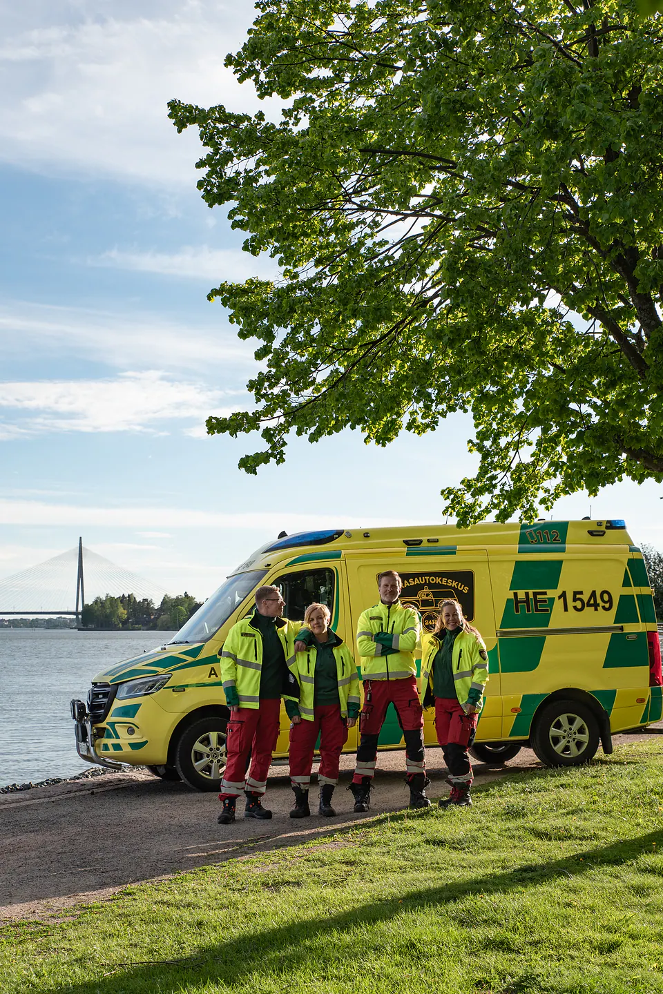 Meidän työyhteisömme Neljä työntekijää seisoo rennossa yhteiskuvassa ambulanssin edessä puistossa vesistö ja silta taustallaan.