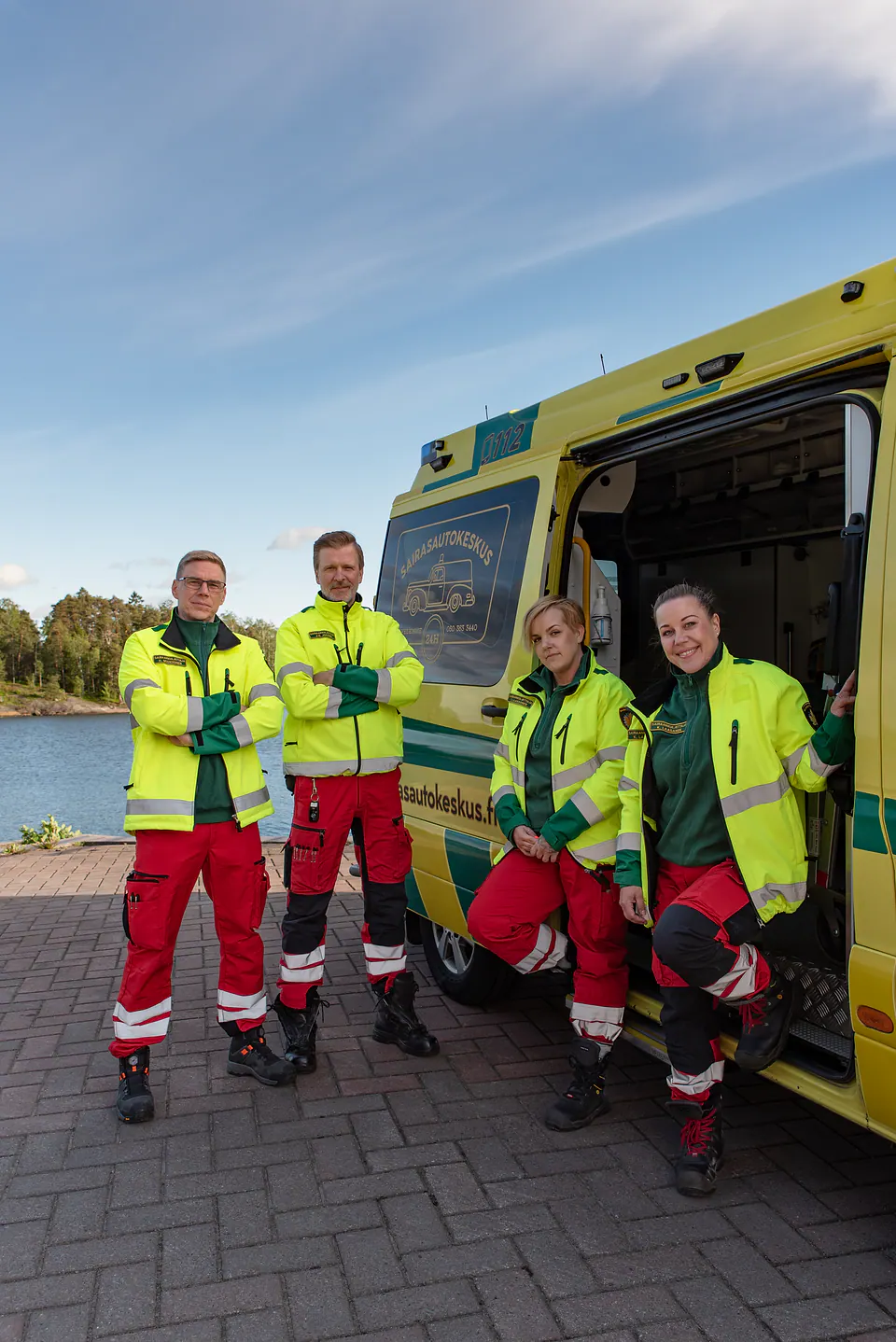 Henkilöstö yhteiskuvassa Neljä työntekijää seisoo kameralle ambulanssin sivuoven luona vesimaisemassa.