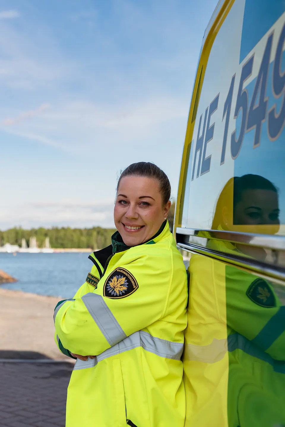 Henkilöstöä rannassa Hymyilevä huomiotakkiin pukeutunut työntekijä nojaa kädet ristissä ambulanssin kylkeen rannan edustalla.