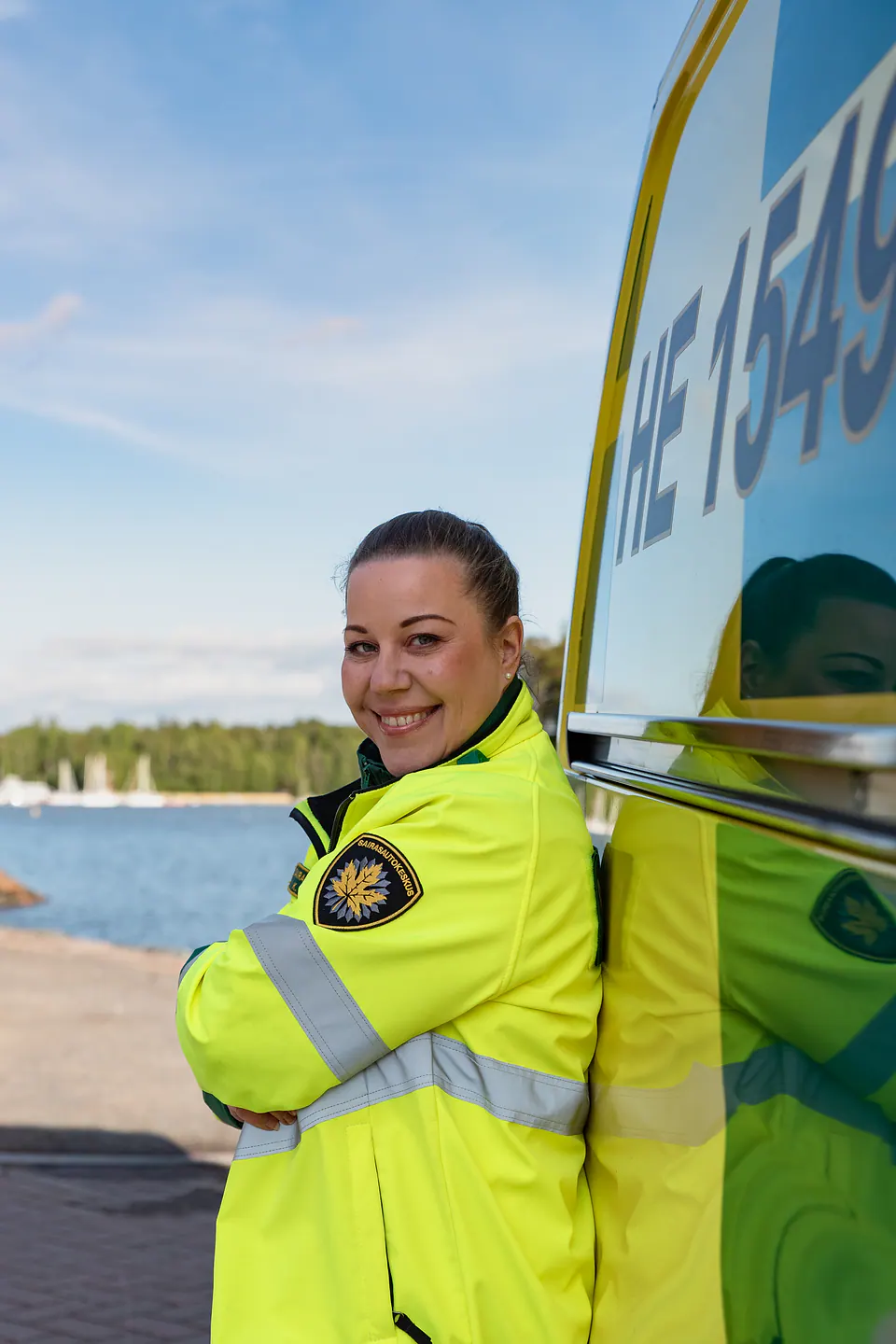 Hymy kaluston äärellä Huomiotakkiin pukeutunut työntekijä nojaa keltaiseen ambulanssiin vesistö taustallaan.