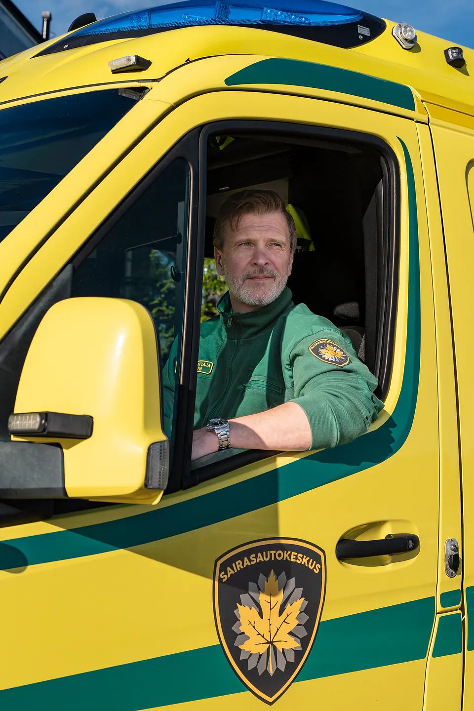 Valmiina ratin taakse Työntekijä istuu keltaisen ambulanssin kuljettajan paikalla avoimen ikkunan ääressä auringonpaisteessa.