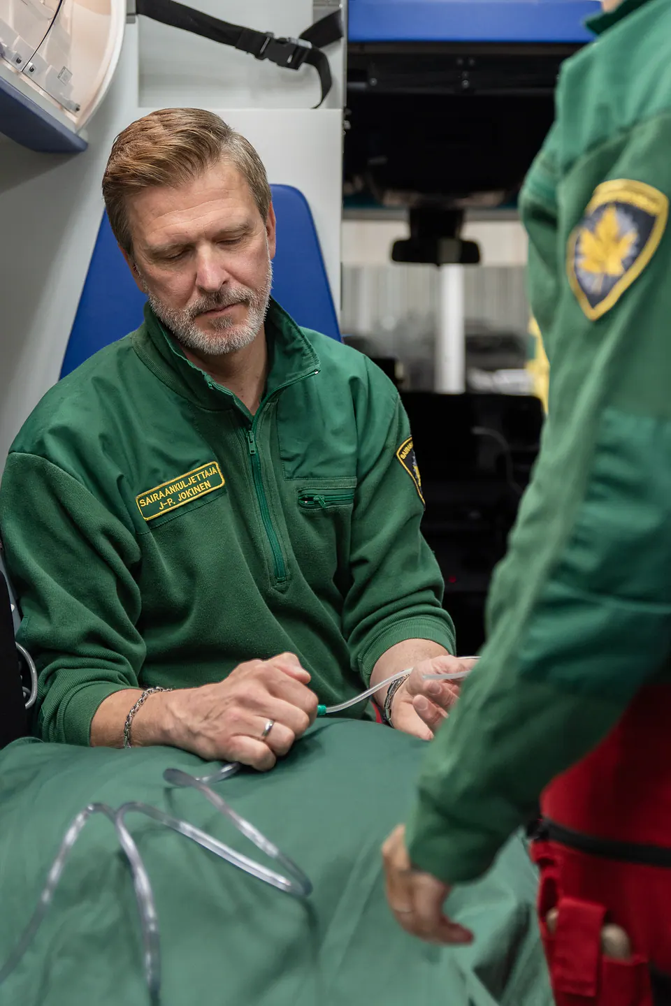 Varusteen yhteinen tarkistus Istuva työntekijä pitelee läpinäkyvää letkua toisen työntekijän seistessä vierellä ambulanssissa.