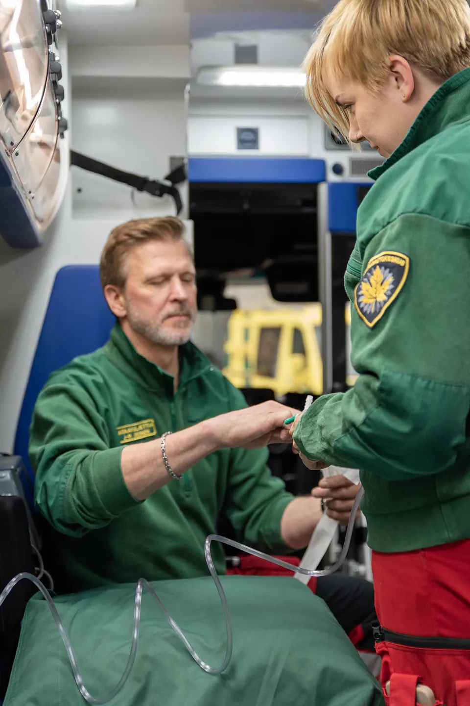 Kädet samassa tehtävässä Kaksi työntekijää kiinnittää yhdessä liitintä läpinäkyvään letkuun ambulanssin sisällä.