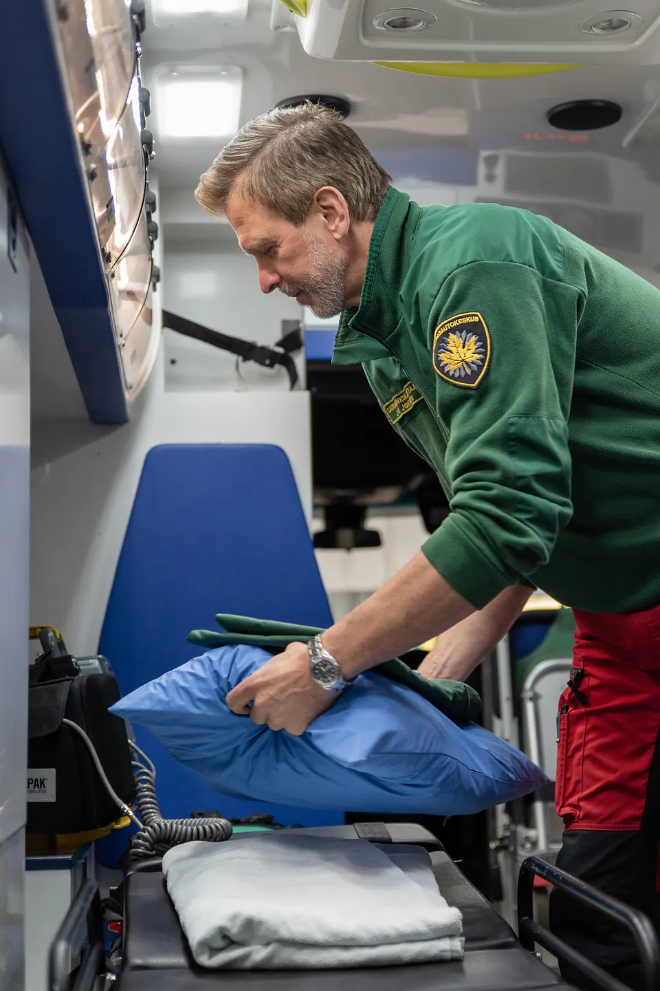 Paarit valmiiksi Työntekijä asettelee sinistä tyynyä ja vihreää peitettä paareille ambulanssin takaosassa.