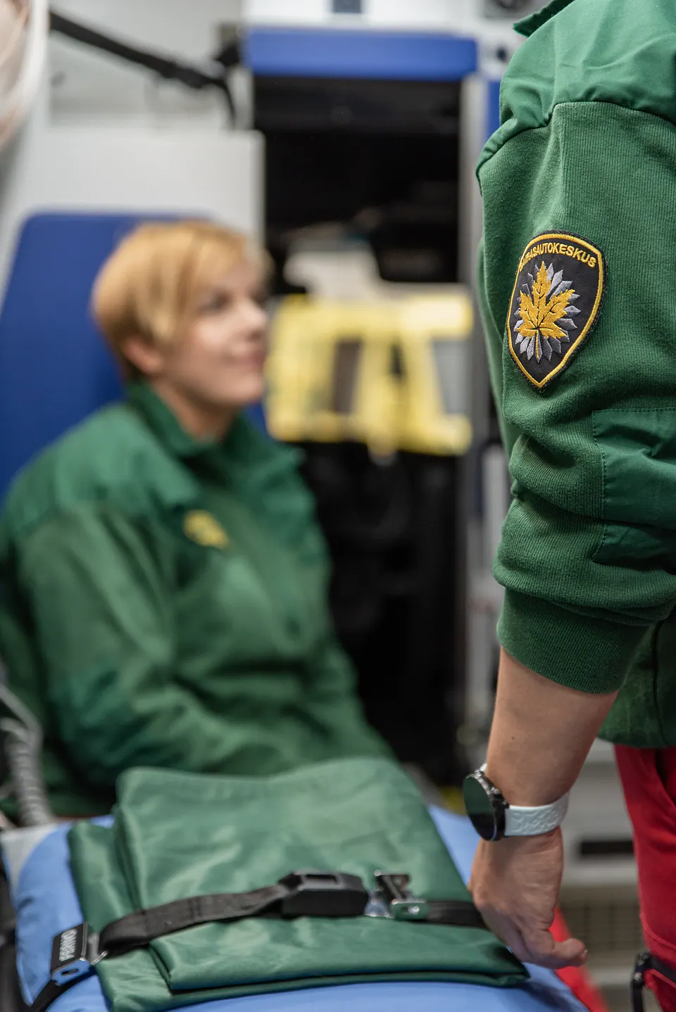 Tunnus työasun hihassa Sairasautokeskuksen hihamerkki näkyy tarkkana etualalla ja työntekijä istuu epäterävänä ambulanssin sisällä.
