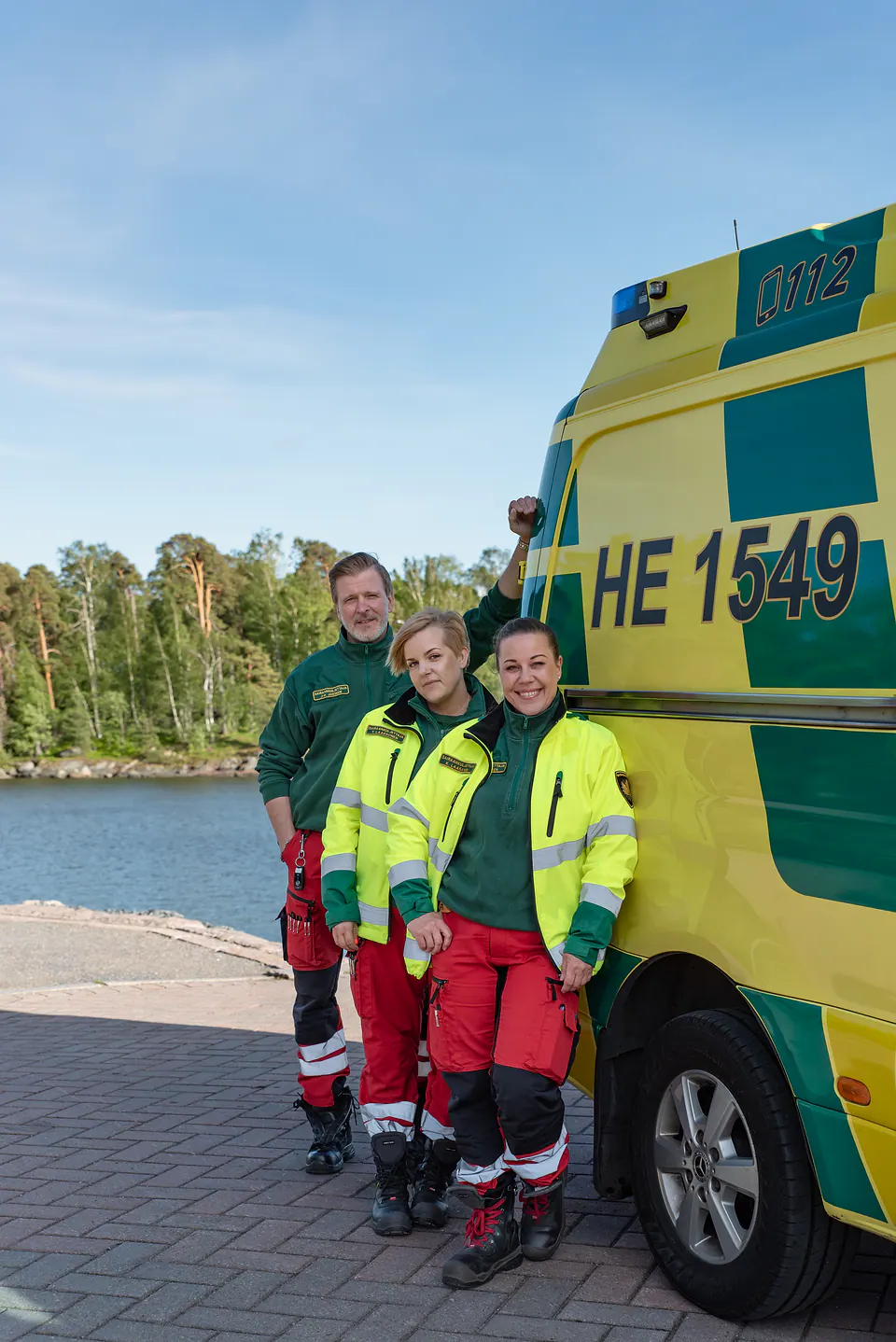Tiimi rannassa Kolme työntekijää seisoo keltaisen ambulanssin vierellä vesistön ja metsän edessä.