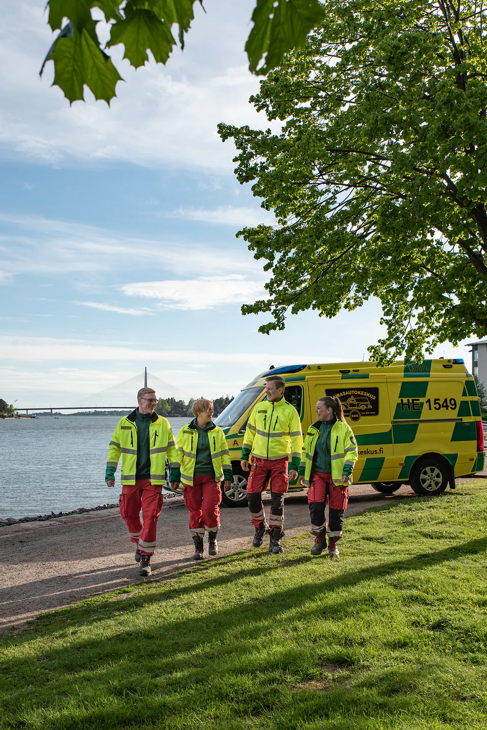 Työyhteisö liikkeessä Neljä työntekijää kävelee keskustellen rantapuistossa ambulanssin vierellä.