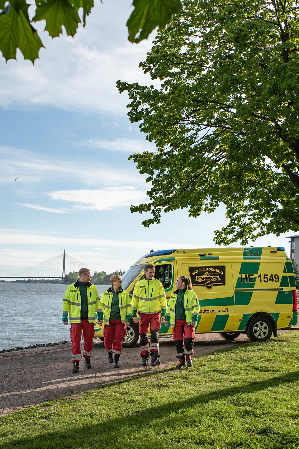 Yhdessä eteenpäin Neljä työntekijää kävelee rinnakkain puistoreitillä ambulanssin ja vesistön edessä.
