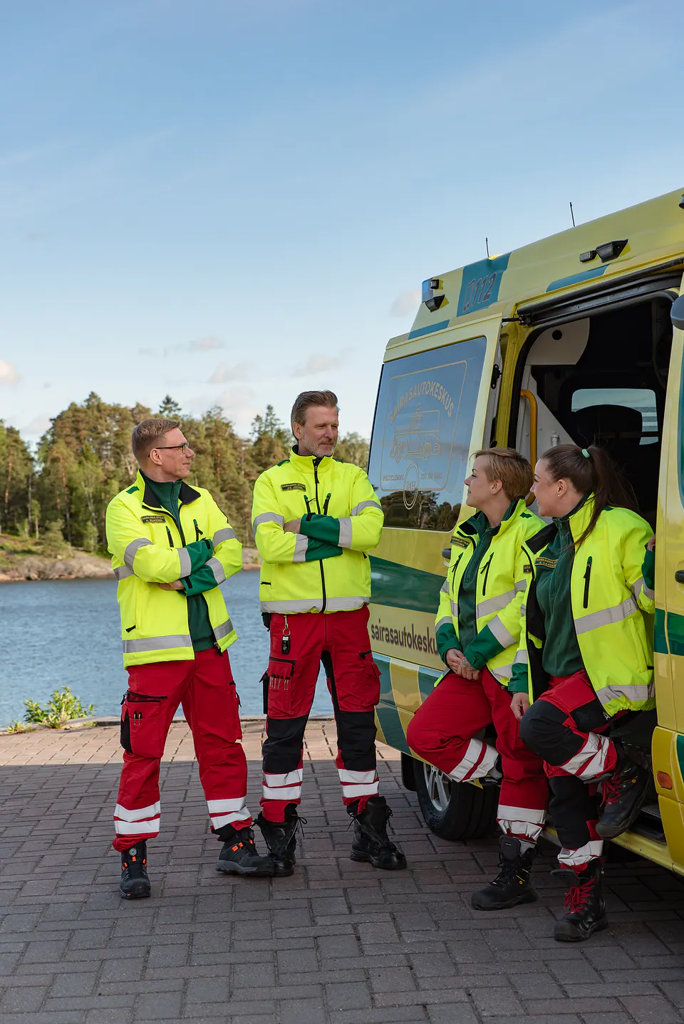 Tiimi yhdessä Neljä työntekijää keskustelee ambulanssin vierellä rannassa, osa seisten ja osa oviaukkoon nojaten.