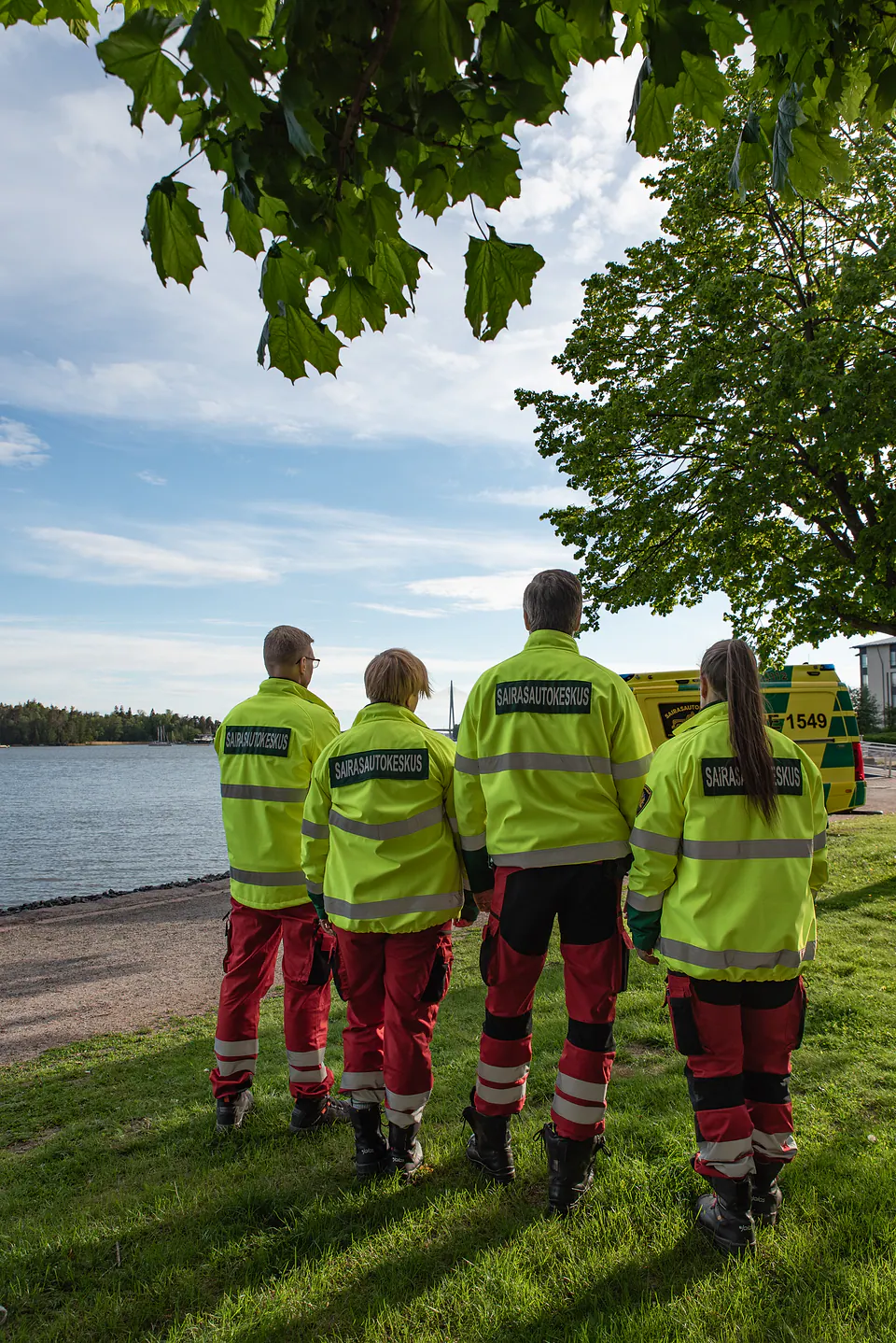 Sama suunta Neljä työntekijää seisoo selin kameralle rannalla, ja ambulanssi näkyy heidän vierellään.