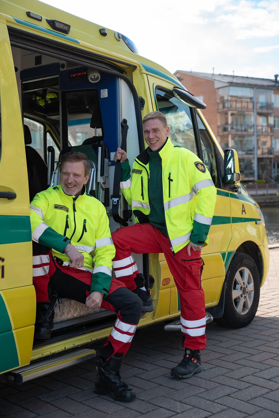 Hyvä hetki työparin kesken Kaksi huomiotakkiin pukeutunutta työntekijää nauraa avoimen ambulanssin ovella kaupunkirannassa.