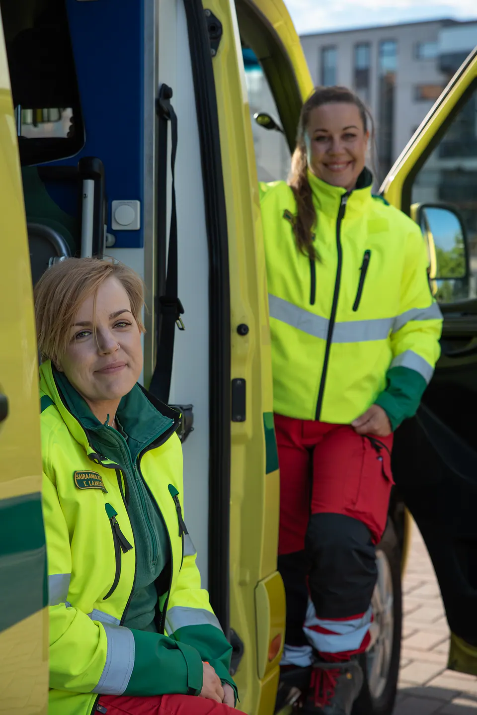 Kaksi työkaveria Kaksi työntekijää on avoimen ambulanssin yhteydessä, etualan henkilö kameraan katsoen ja taustan henkilö hymyillen.