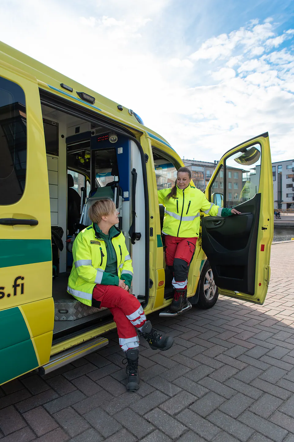 Yhdessä auton äärellä Kaksi työntekijää on avoimen ambulanssin ovilla, toinen istuen sivuovella ja toinen seisten etuovella.