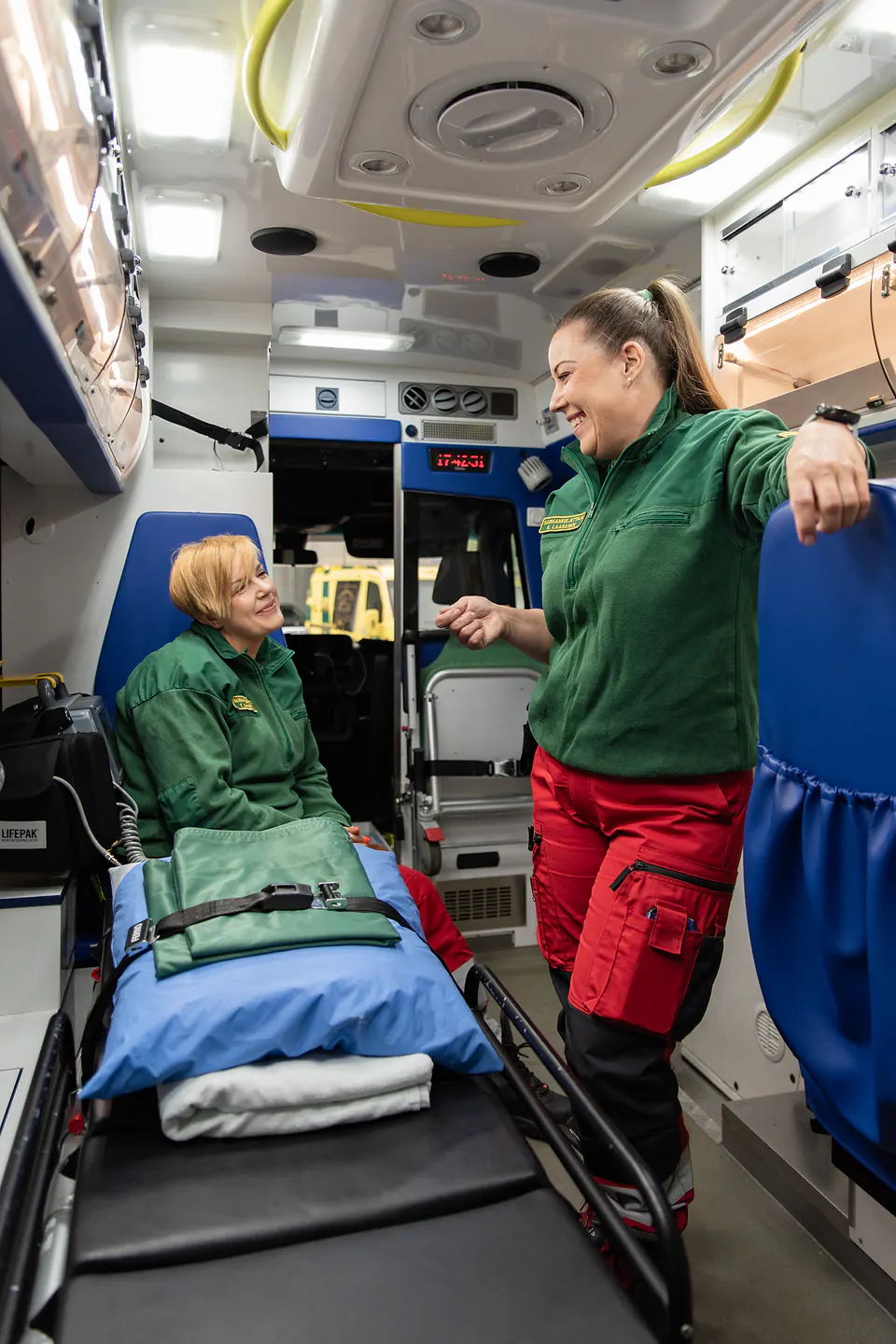 Rento hetki työparin kesken Kaksi työntekijää hymyilee ja keskustelee ambulanssin takaosassa paarien vierellä.