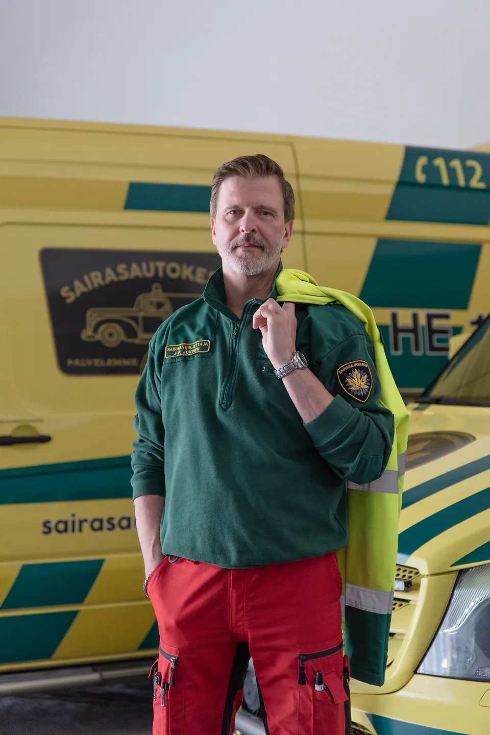 Ammattilainen kaluston äärellä Työntekijä seisoo ambulanssihallissa huomiotakki olallaan kahden keltaisen ambulanssin edessä.