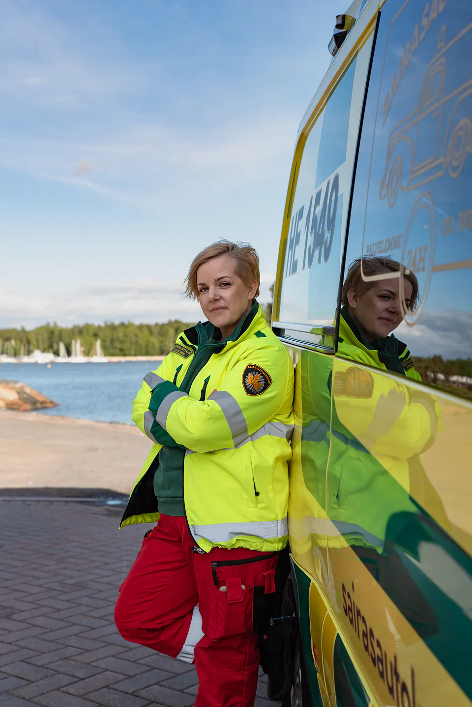 Työntekijä ja ambulanssi Huomiotakkiin pukeutunut työntekijä seisoo kädet ristissä ambulanssin kyljellä vesimaisemassa.