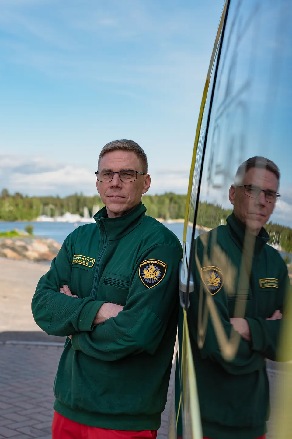 Henkilöstömuotokuva Silmälasipäinen työntekijä seisoo kädet ristissä ambulanssin vierellä heijastuen auton kylkeen.