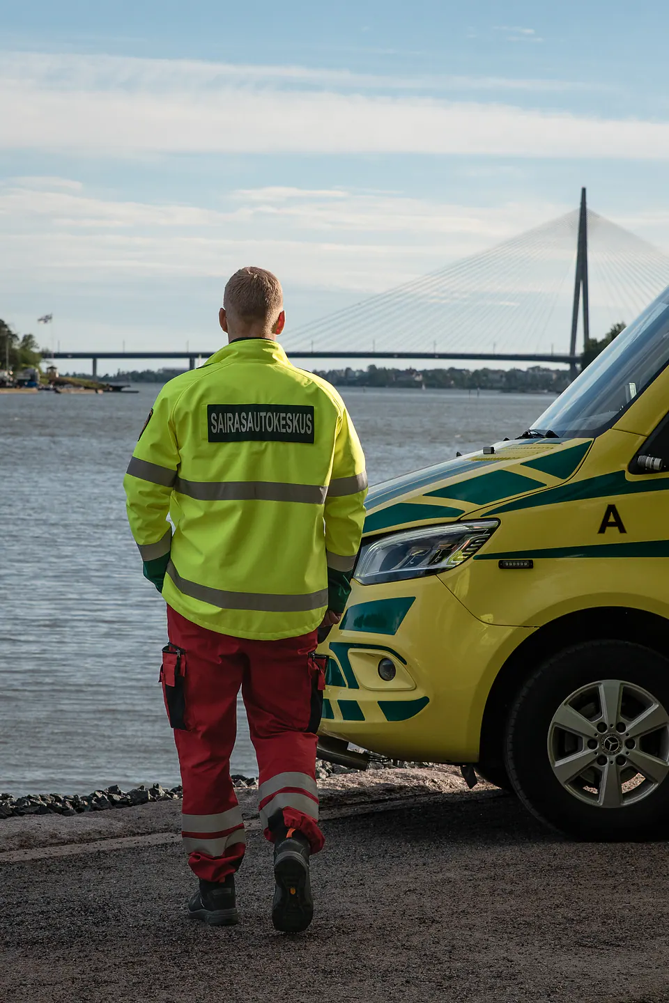 Kohti autoa Selin kuvattu työntekijä kävelee kohti rantaan pysäköityä ambulanssia vinoköysisilta taustallaan.