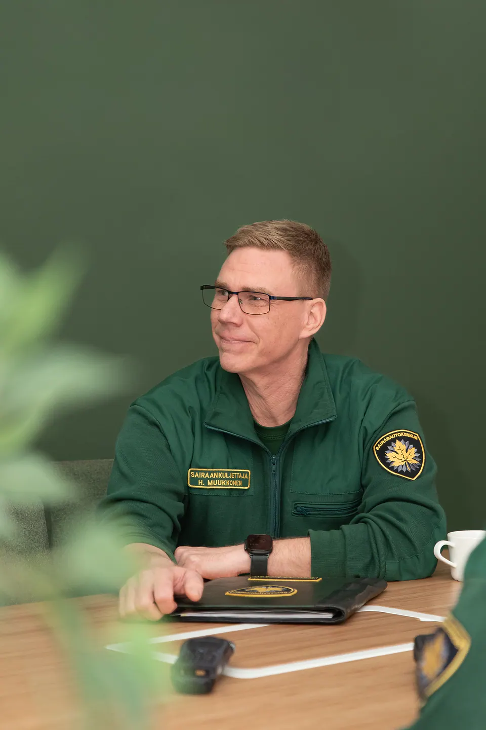 Keskittyneenä yhteiseen hetkeen Vihreään työasuun pukeutunut työntekijä istuu palaveripöydän ääressä kansio edessään.