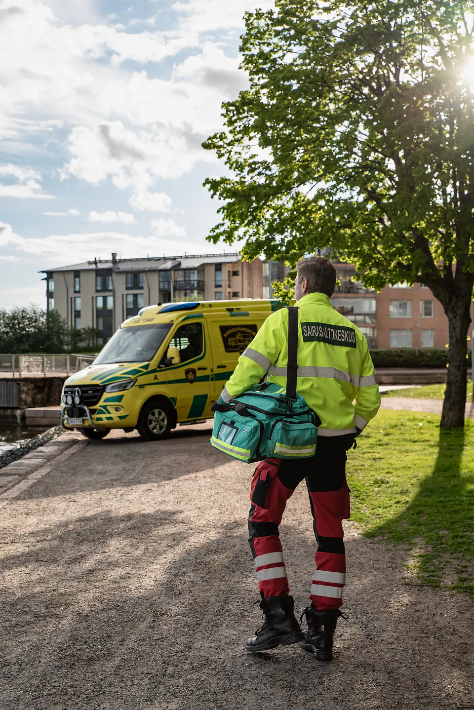 Valmiina lähtöön Työntekijä seisoo selin kameraan varustelaukku olallaan ja katsoo rantareitillä olevaa ambulanssia.