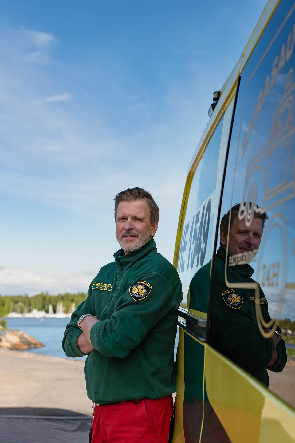Katse kameralle Työntekijä seisoo kädet ristissä ambulanssin kyljellä, ja hänen heijastuksensa näkyy auton pinnassa.