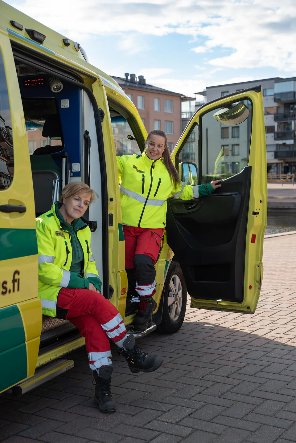Työpari valmiina päivään Kaksi työntekijää katsoo kameraan ambulanssin avointen ovien äärellä, toinen istuen ja toinen seisten.