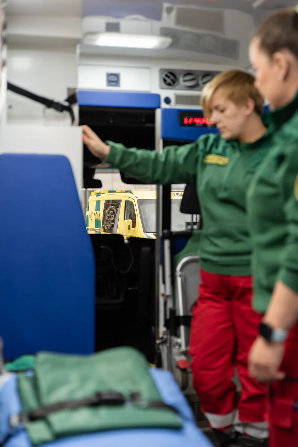Työpari auton sisällä Kaksi vihreään työasuun pukeutunutta työntekijää seisoo ambulanssin takaosassa paarien vierellä.
