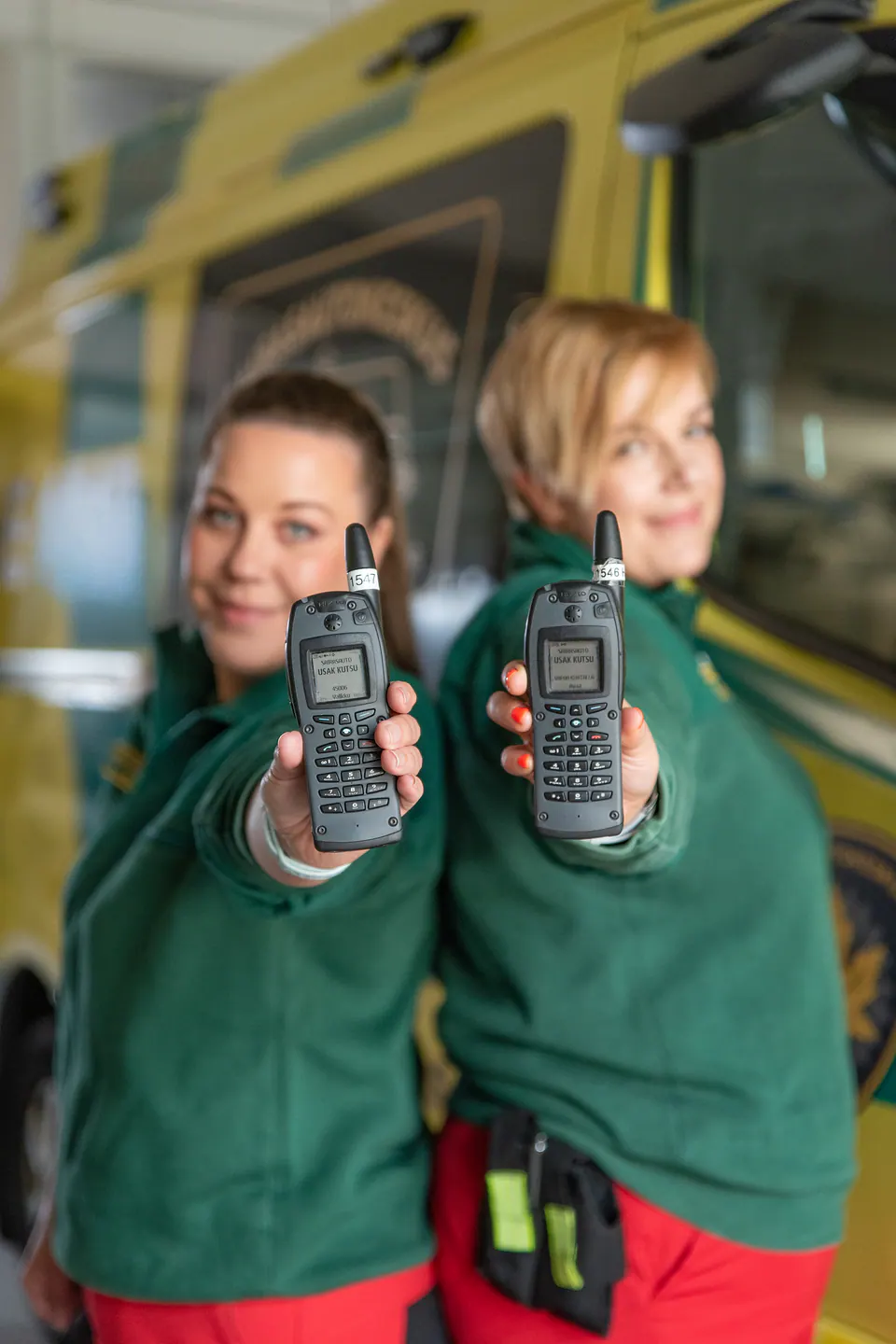 Yhteinen kanava Kaksi työntekijää pitää radiopuhelimia kameran edessä ambulanssin vierellä.