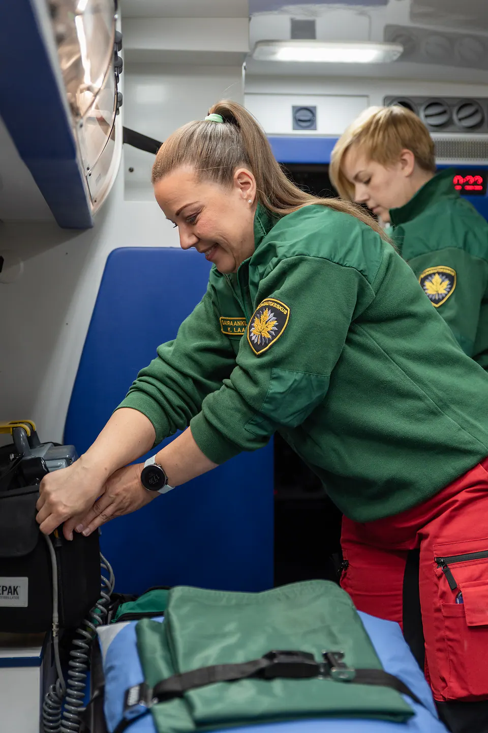 Valmistelu tehdään yhdessä Kaksi työntekijää valmistelee laitetta ja kaapeleita ambulanssin takaosassa paarien vierellä.