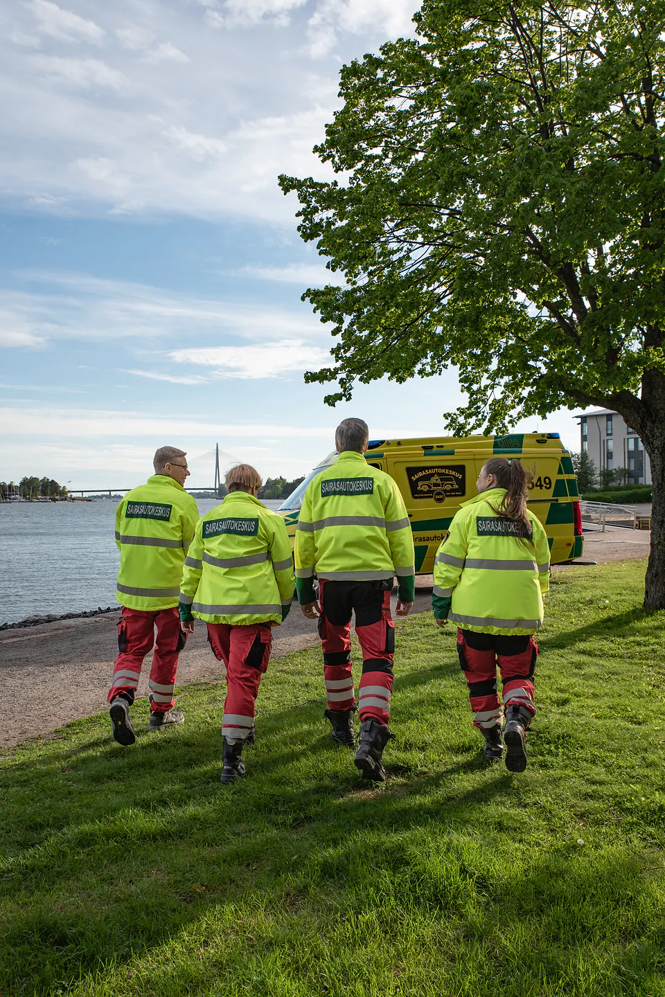 Tiimi matkalla autolle Neljä työntekijää kävelee selin kameralle nurmen halki kohti rantaan pysäköityä ambulanssia.