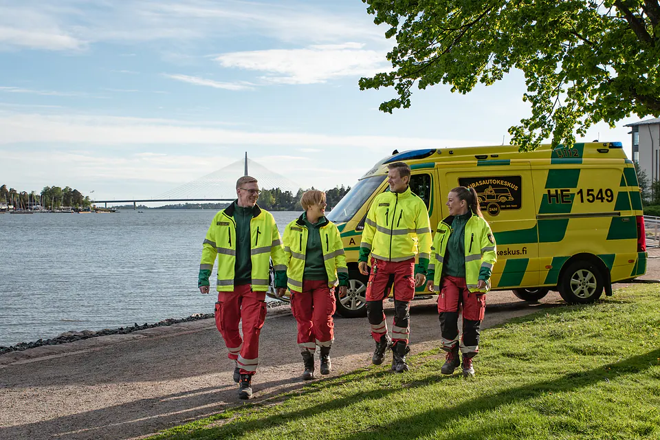 Tiimi rannalla Neljä työntekijää kävelee rinnakkain rantareitillä ambulanssin, vesistön ja sillan edessä.
