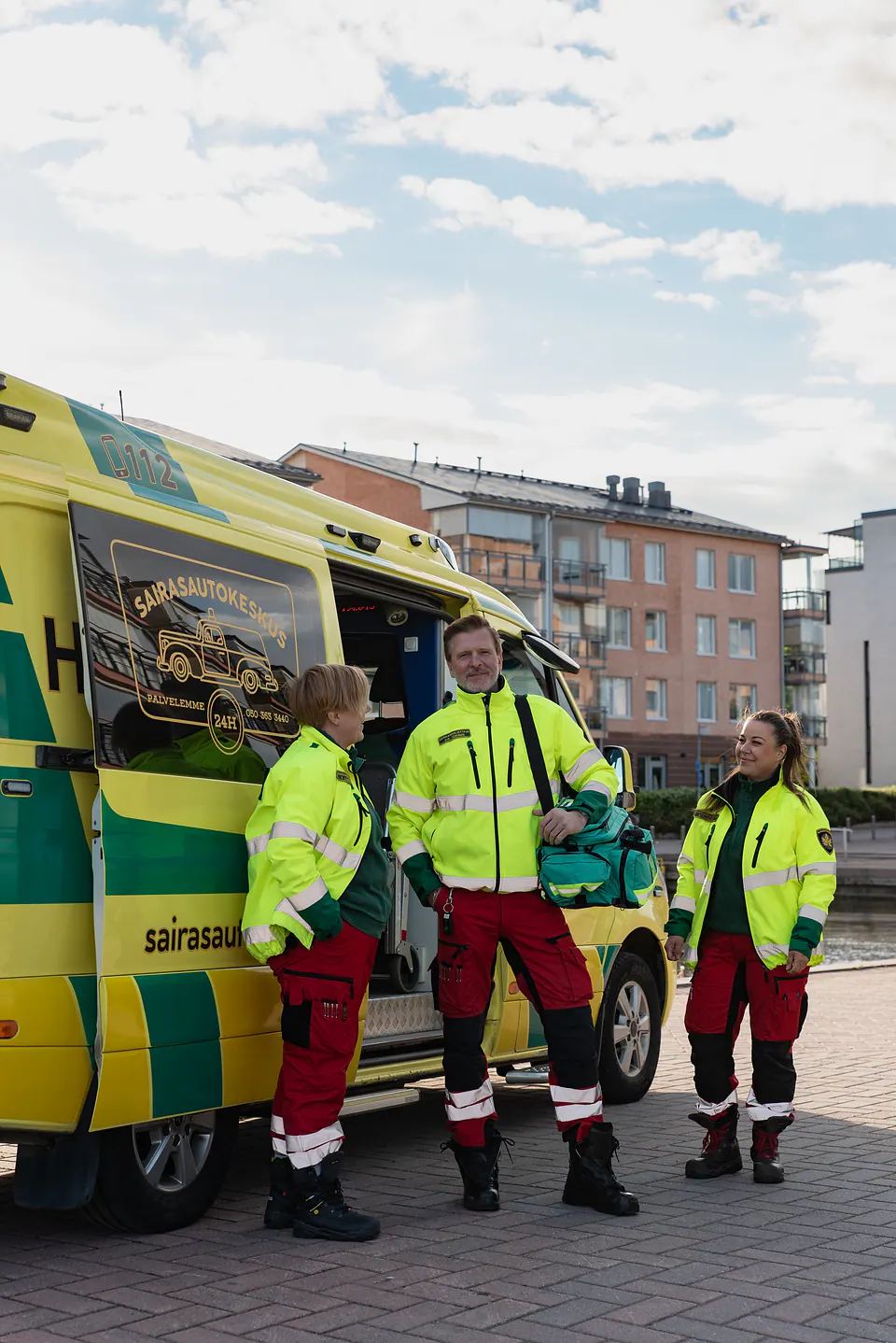 Varusteet mukana Kolme työntekijää seisoo ambulanssin luona, ja keskimmäisellä on vihreä varustelaukku olallaan.