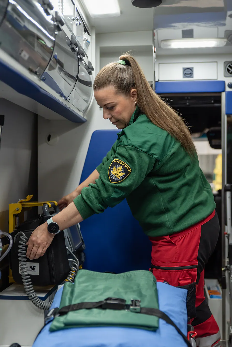 Varusteet järjestykseen Työntekijä tarkistaa laitetta ja sen kaapelia ambulanssin takaosassa paarien vierellä.