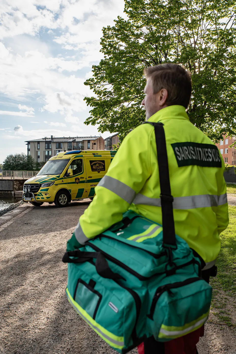Varusteet mukaan Selin kuvattu työntekijä kantaa suurta vihreää varustelaukkua kohti kauempana olevaa ambulanssia.