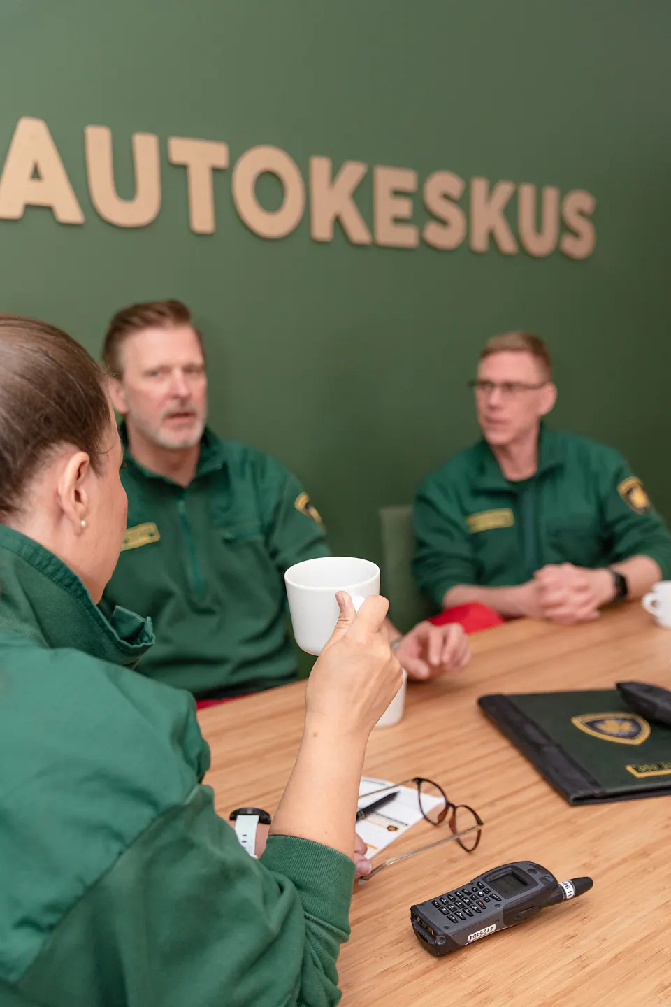 Työpäivän yhteinen tilannekuva Kolme vihreisiin työasuihin pukeutunutta työntekijää keskustelee pöydän ääressä radiopuhelin ja kahvikupit edessään.